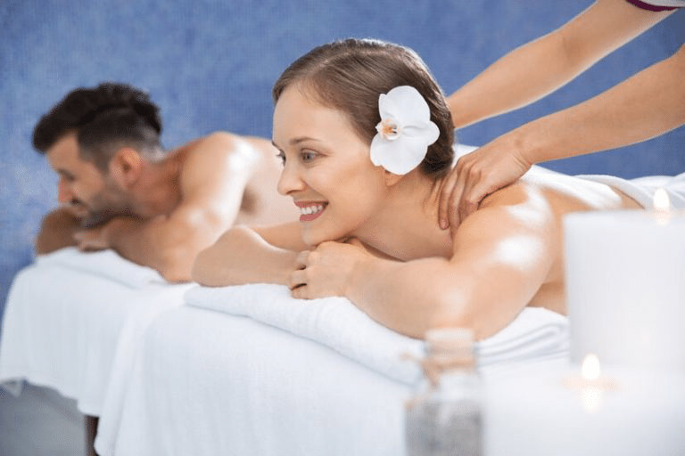 A woman with a flower in her hair enjoys a back massage while lying next to her partner—a shared relaxation and bonding activity.