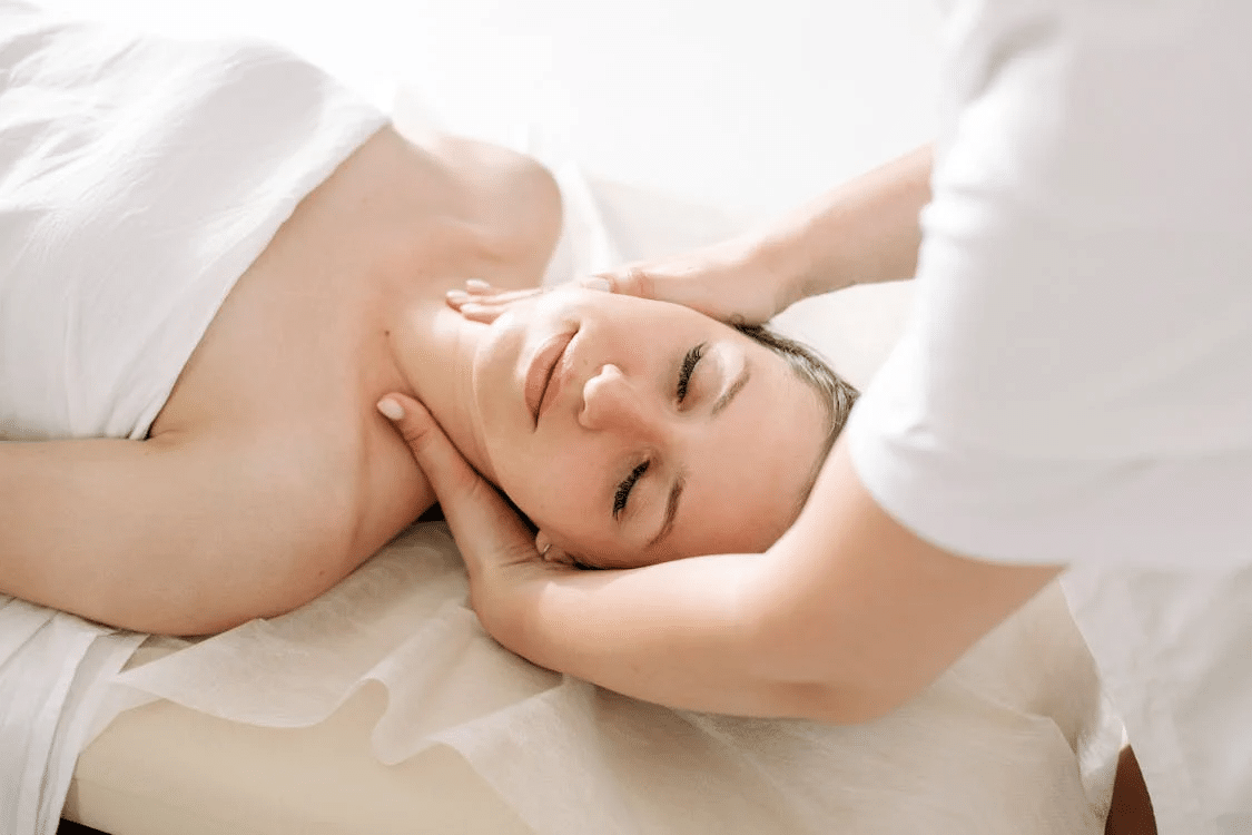 a woman in a white towel on a massage chair getting a neck massage from a masseuse